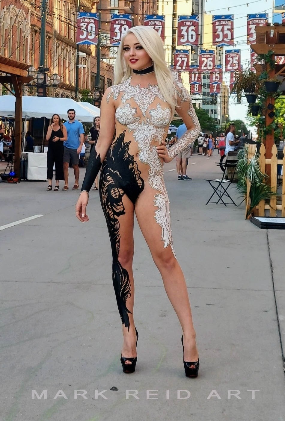 Full body painting on a thin, blonde, long haired woman. She has lace that is white on the her top and trailing down her left leg.  Black lace starting on her right torso under her arm that trails down her right leg.