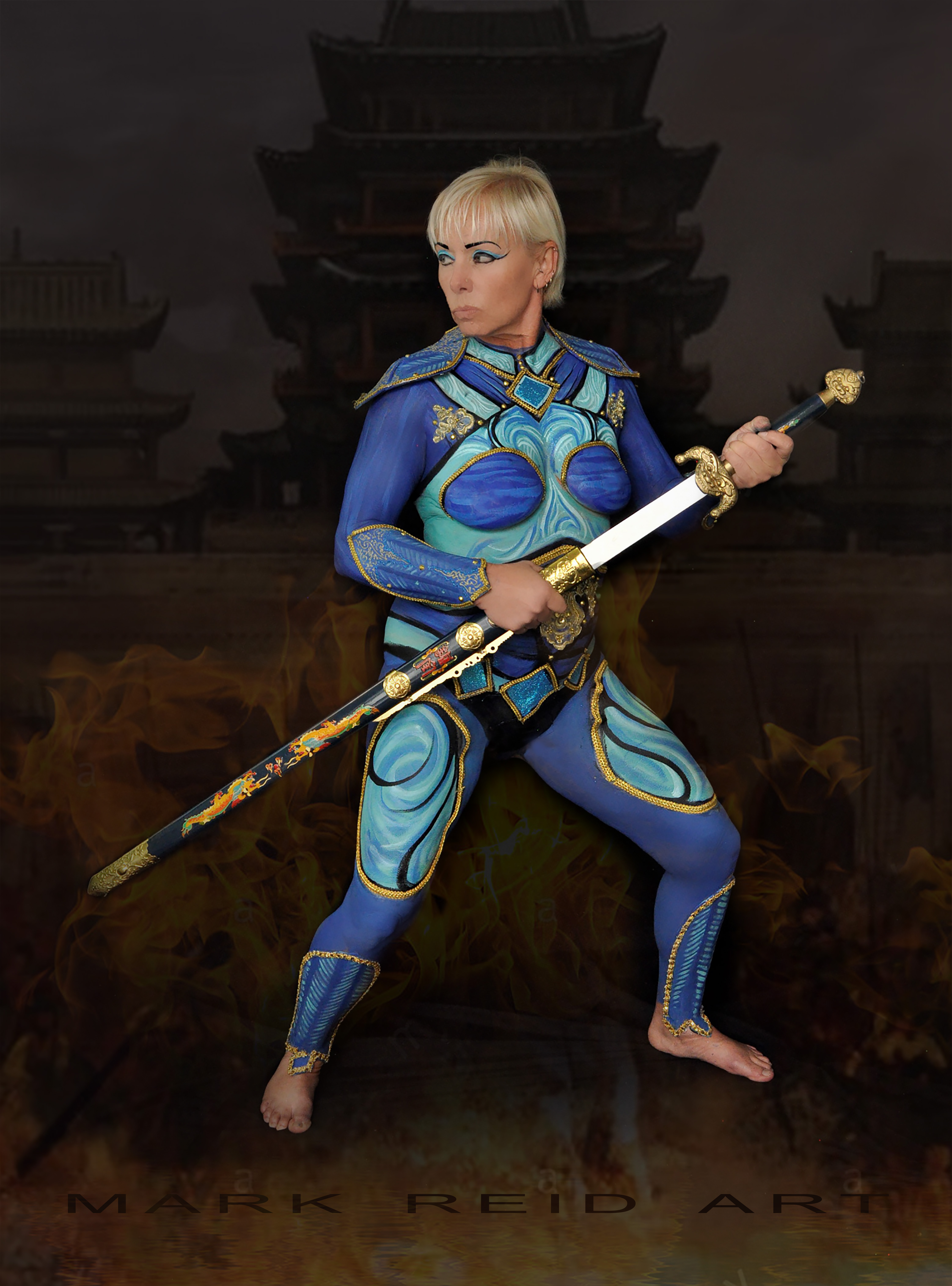 A blonde woman in full body paint that looks like blue and turqoise armor. She is holding a sword.