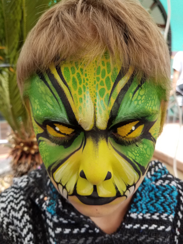 boy in a face painting that is a green and yellow lizzard face featuring snake eyes when his eyes are closed