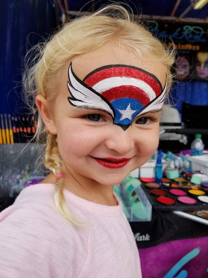 Young girl with a painting of wings above her eyes with a red white and blue pattern on her forehead with a white star between her eyes