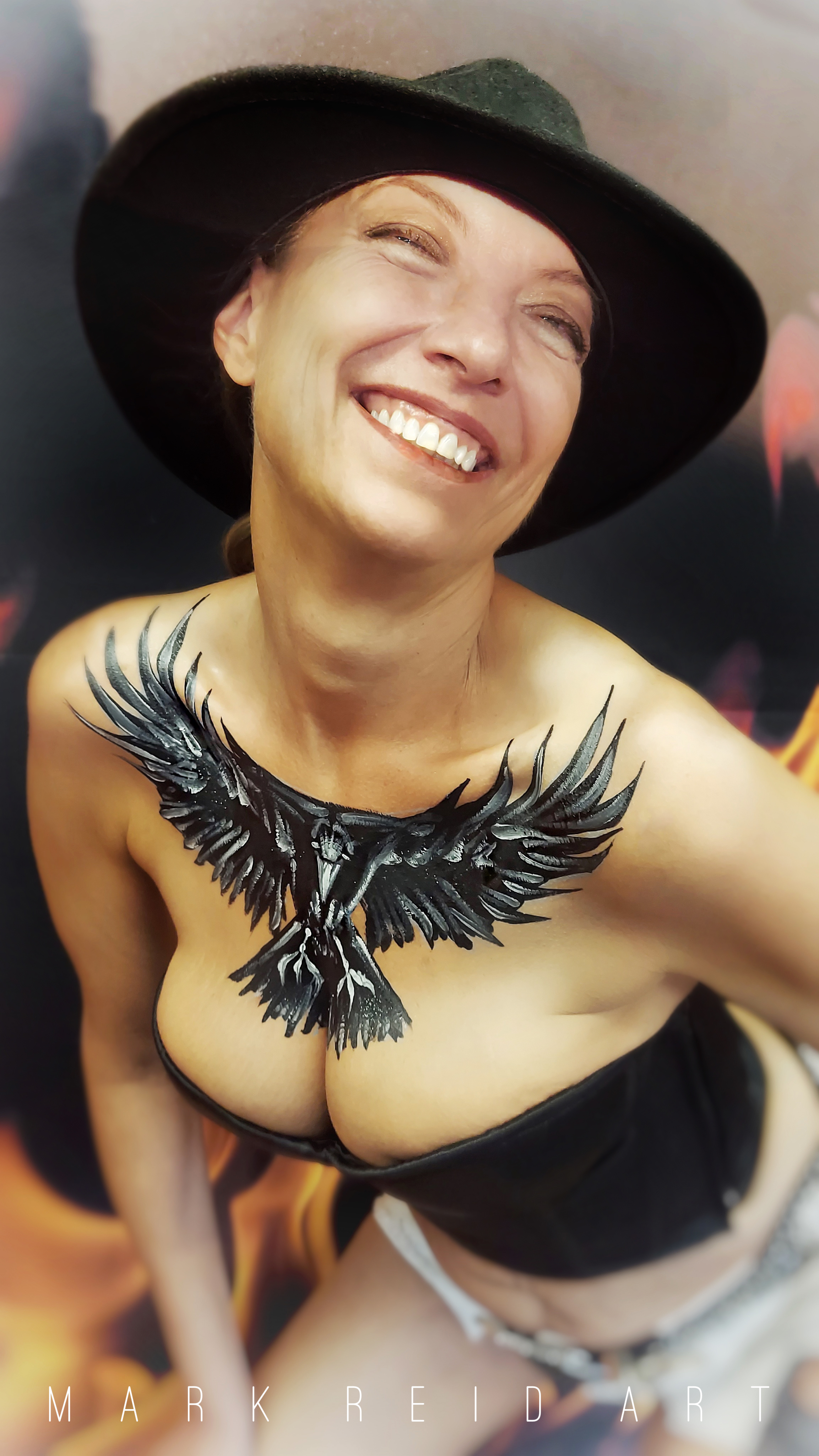 Smiling woman wearing a black fedora with an extremely detailed black and white eagle in flight painted on her upper chest with the feathers reaching up to her callar bones and out to the the shoulders 