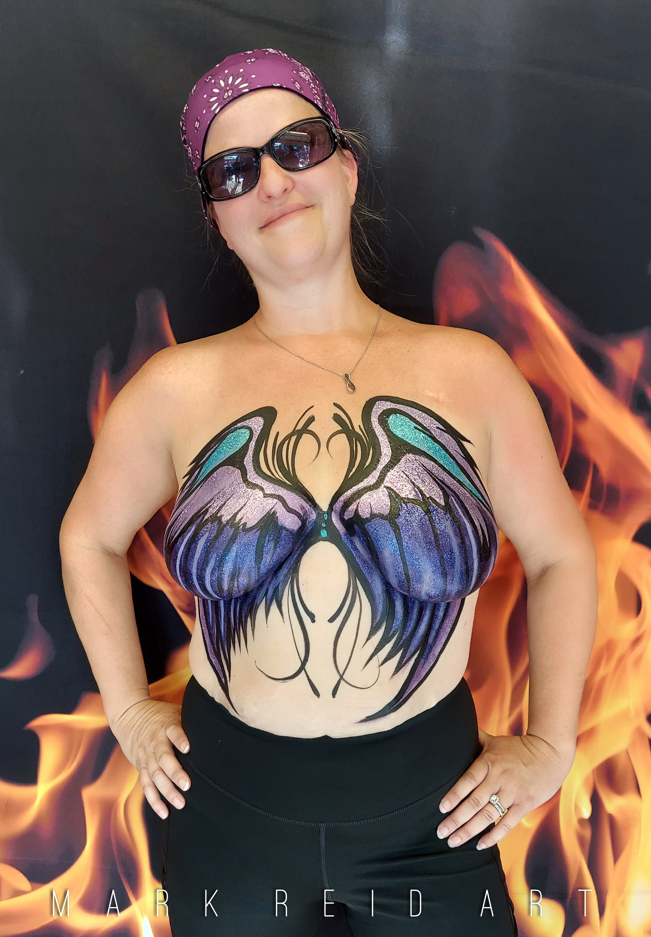 Dark haired woman with a puprle headband sporting a painted on top of light blue, medium blue, and a light purple. The wings originate from her sternum and spread across her breasts with finely detailed feathers that reach down to her waistline