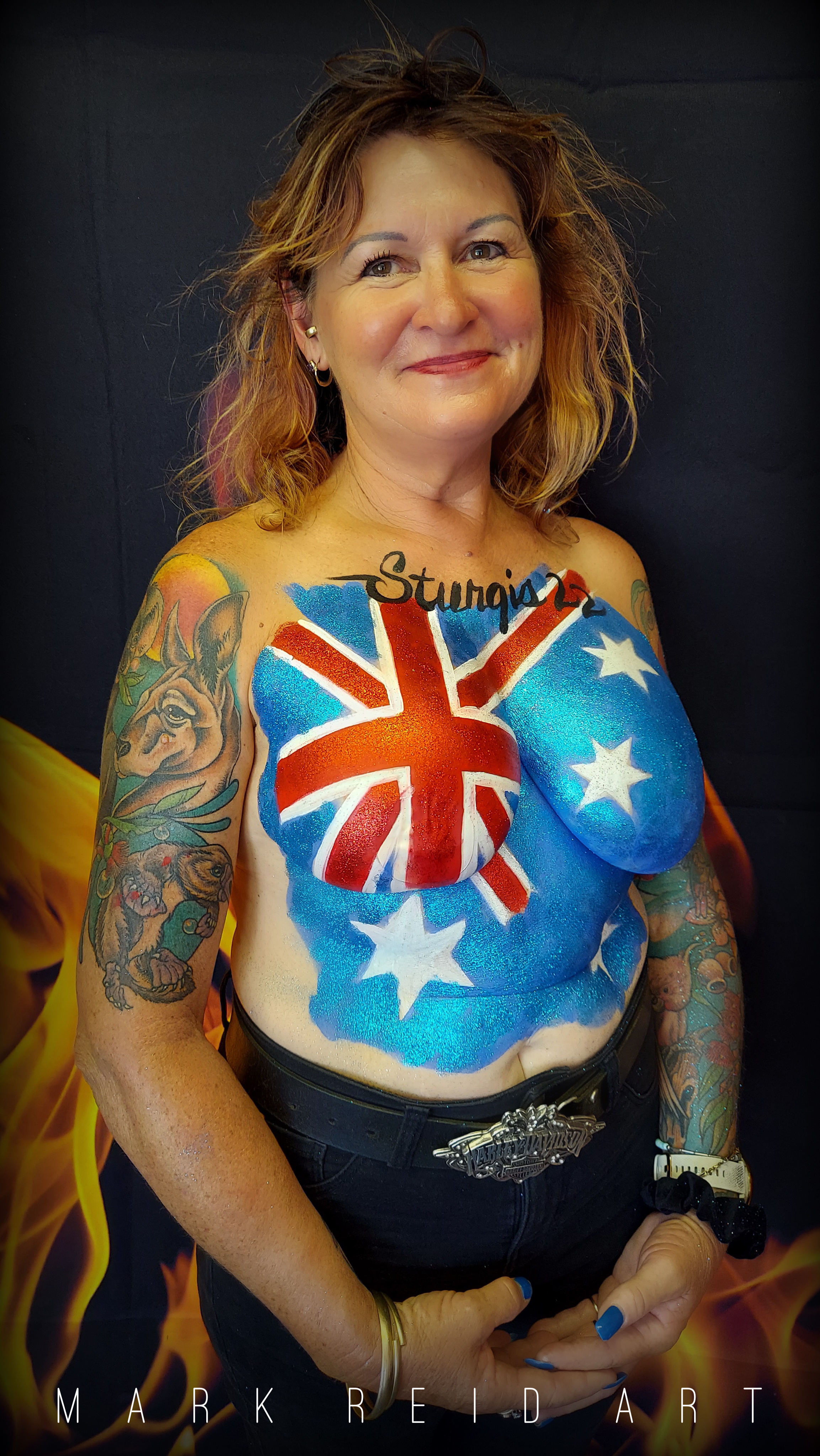 Redish-blonde haired woman with a painted on top that is the Australian flag across her chest with the word Sturgis at the top center near her collar bone