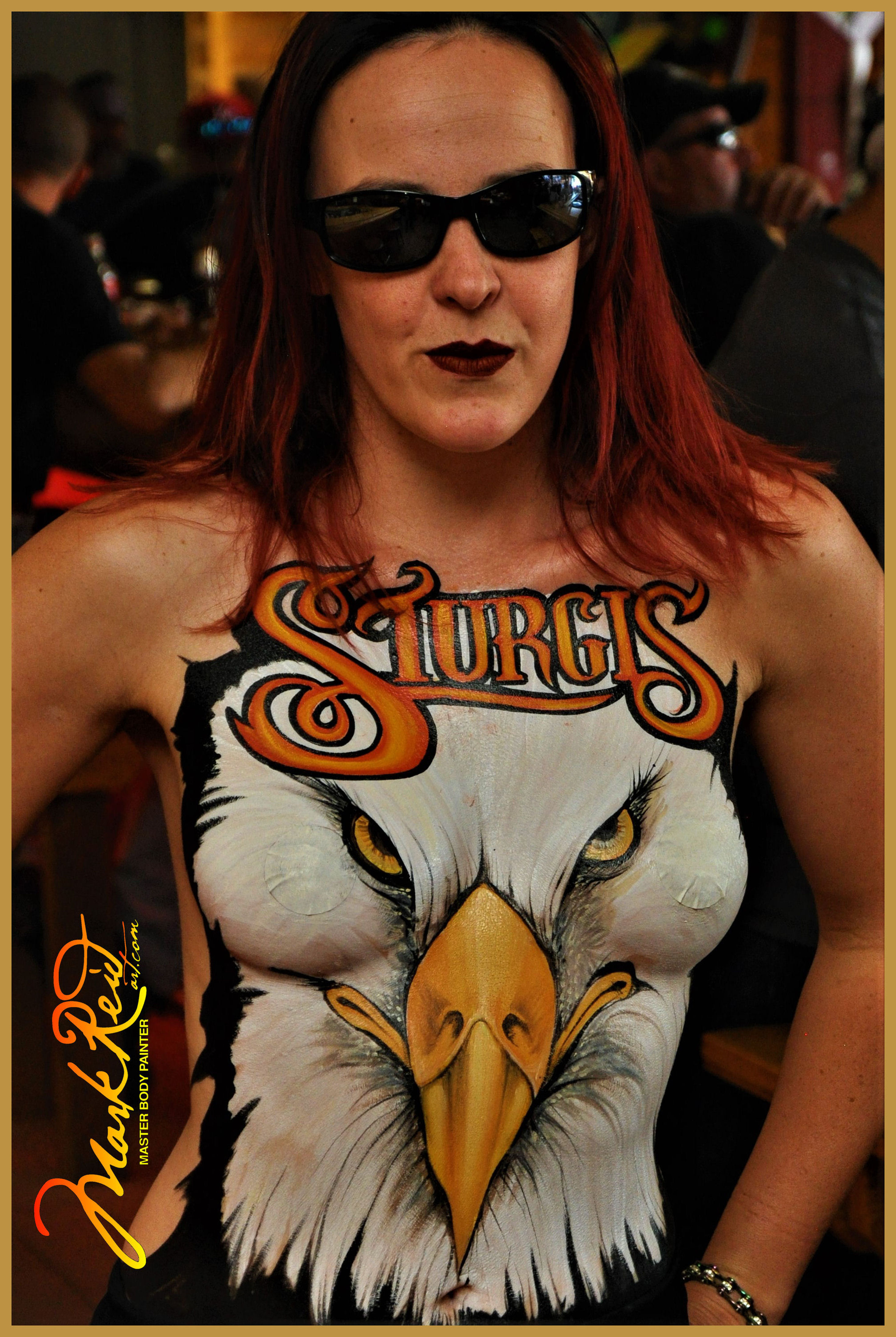 model with long red hair with a body painting of a close up of an eagle's face on her chest and the word Sturgis above it